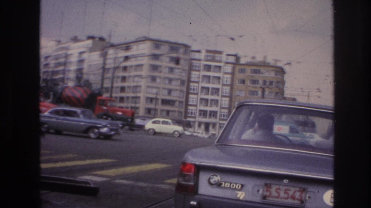 1967 Paris street view from inside a vehicle, showing a grey BMW 1800 near a crosswalk with traffic and apartment buildings behind it.