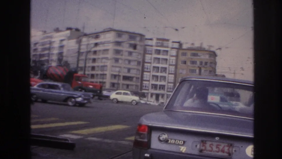 1967 Paris street view from inside a vehicle, showing a grey BMW 1800 near a crosswalk with traffic and apartment buildings behind it.