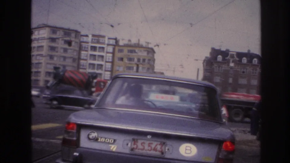 1967 Paris road scene showing the rear of a grey BMW, surrounding traffic, a red cement mixer, and apartment buildings from an in-car viewpoint.