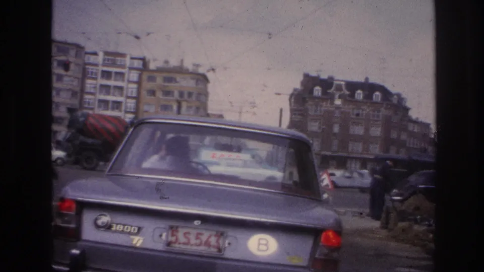 1967 Paris street footage with a grey BMW in the foreground, roadwork activity to the right, and city traffic around apartment buildings.