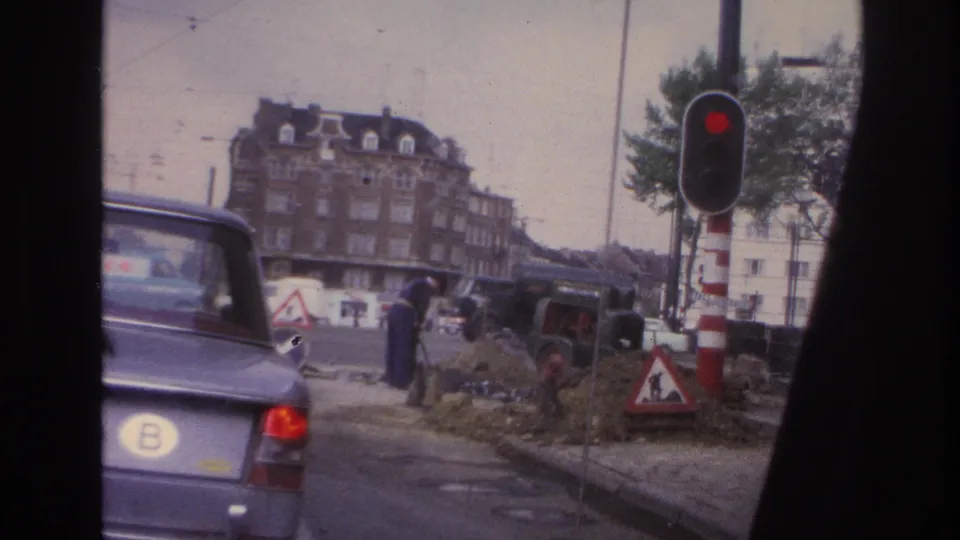 Paris street in 1967 with a red signal, roadwork signs, a worker, a green truck, and the rear of a blue BMW at frame edge.