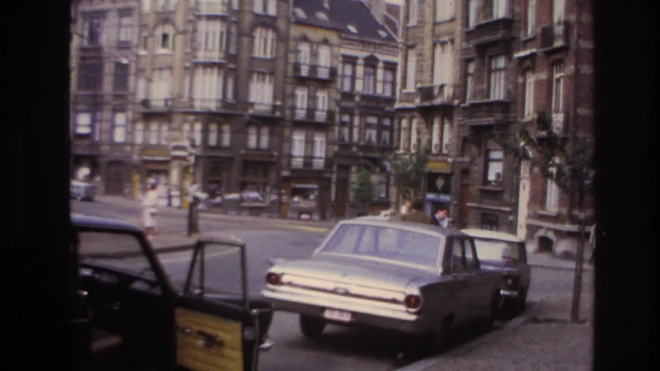 1967 Paris street scene with a parked sedan, open vehicle door, pedestrians crossing or walking, and dense residential facades.