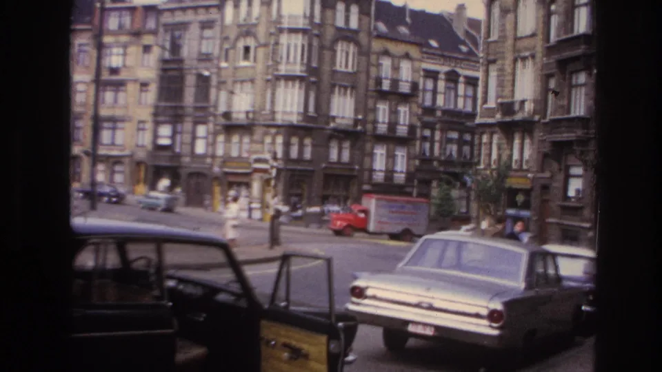 1967 Paris street with a light sedan, dark car with open door, red delivery truck, pedestrians, and older apartment facades.