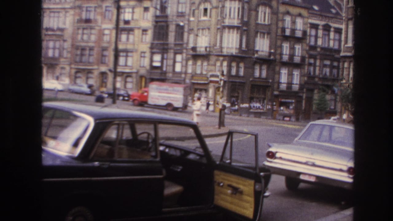 1967 Paris street scene with a dark foreground car, open door, light sedan, delivery truck, and ornate apartment buildings.