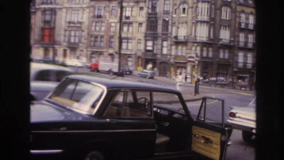 1967 Paris vehicle-side view showing a black car, open door, parked cars, pedestrians, and tall apartment facades.