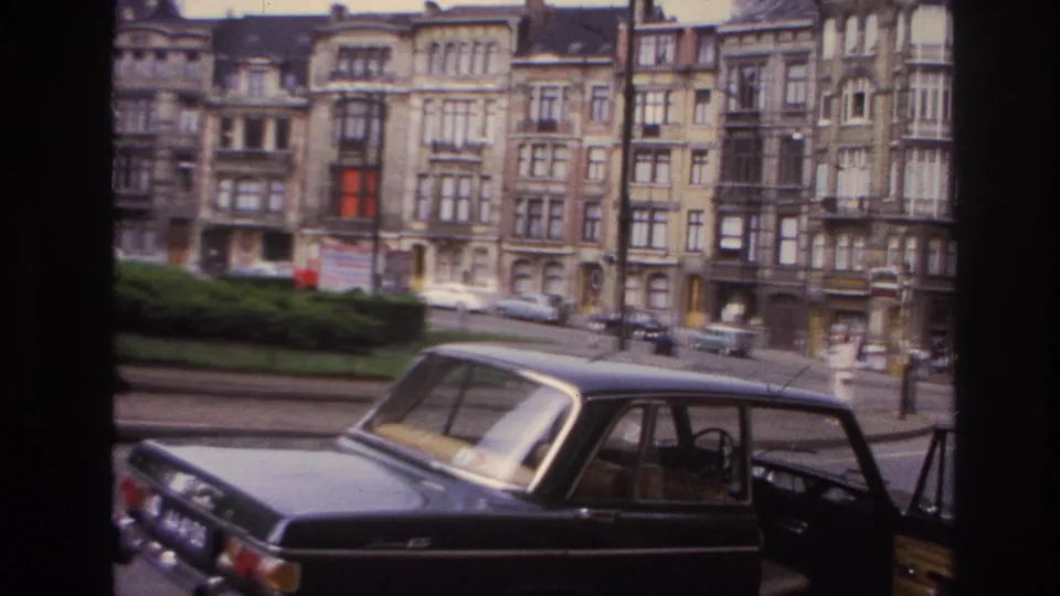 1967 Paris street view from 8mm footage showing a black sedan in the foreground and rows of ornate apartment buildings beyond.