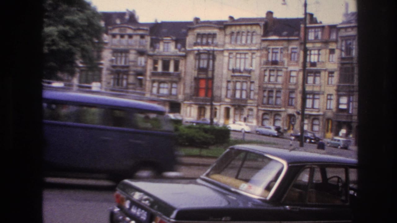 Blue van passing a black sedan with apartment buildings and hedges in the background.