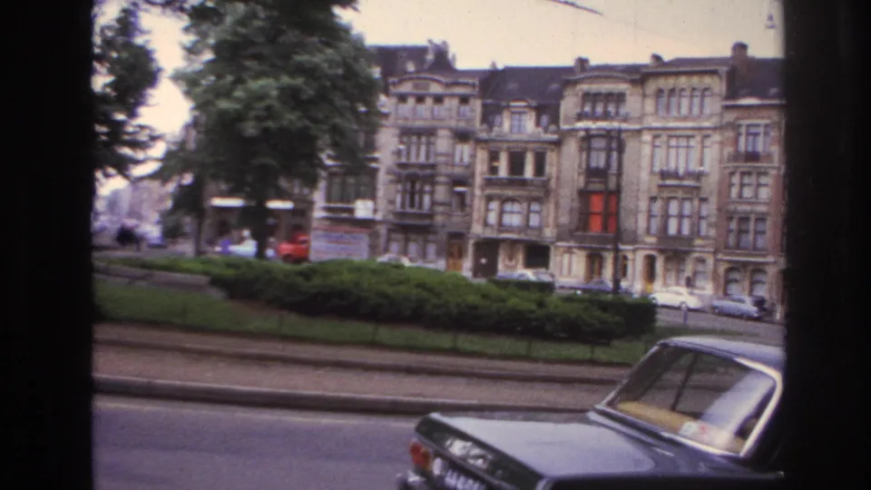 Moving 8mm view of a Paris street edge with hedges, mature trees, parked cars, and ornate apartment buildings.