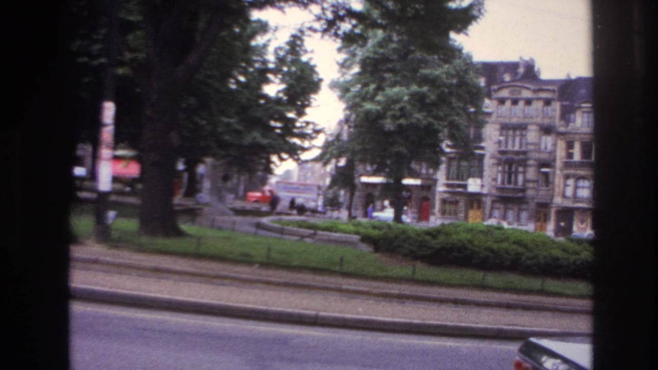 1967 Paris home-movie frame showing a tree-lined city street, passing cars, ornate buildings, and dark vehicle-window framing.