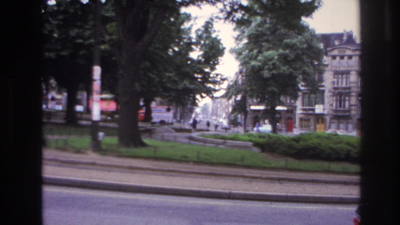 1967 Paris street view from a moving vehicle, framed by dark window edges with cars, trees, and distant pedestrians.