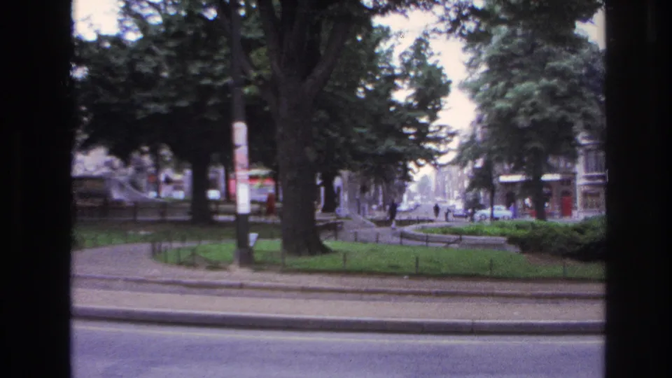 1967 Paris frame with a large foreground tree, planted median, roadway, distant traffic, and pedestrians in soft 8mm focus.