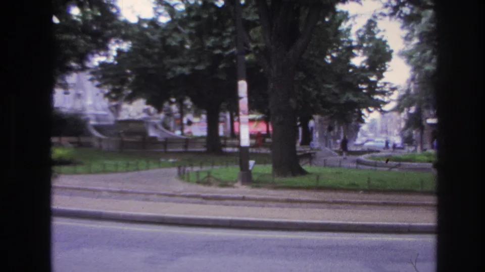 1967 Paris home-movie frame with motion blur, a foreground tree, street median, passing traffic, and distant urban facades.