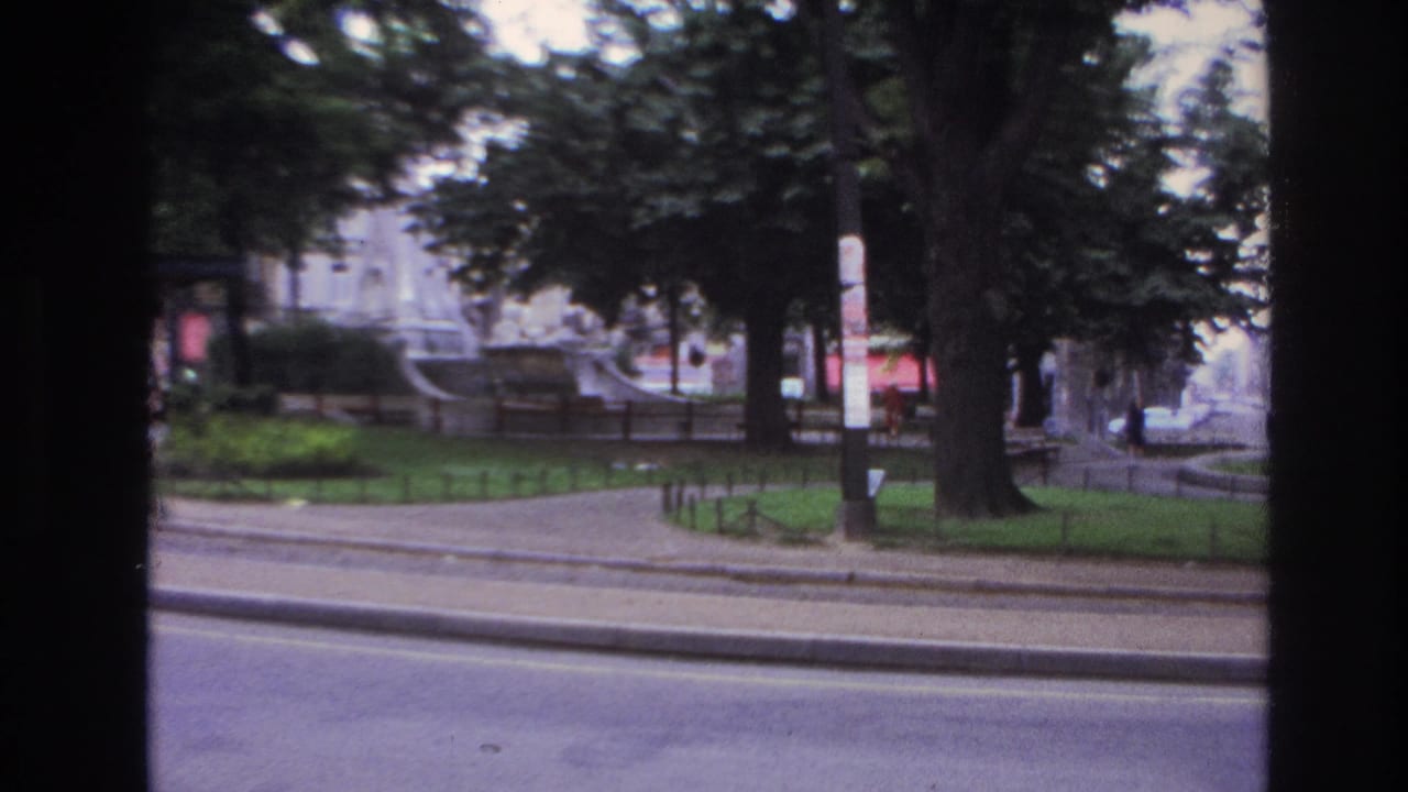 1967 Paris home-movie frame showing a park, fountain, kiosk, trees, sidewalk railings, and street viewed through dark film-frame edges.