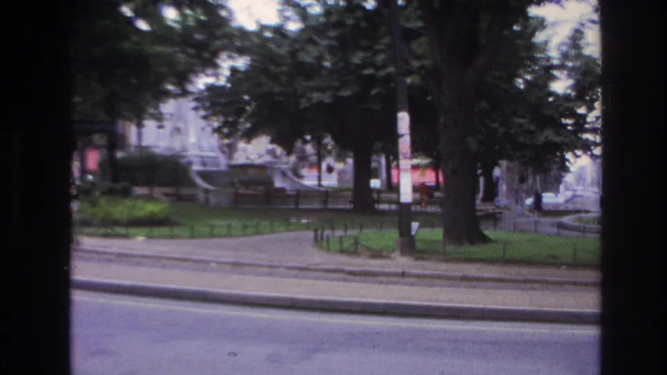 1967 Paris home-movie frame showing a park, fountain, kiosk, trees, sidewalk railings, and street viewed through dark film-frame edges.