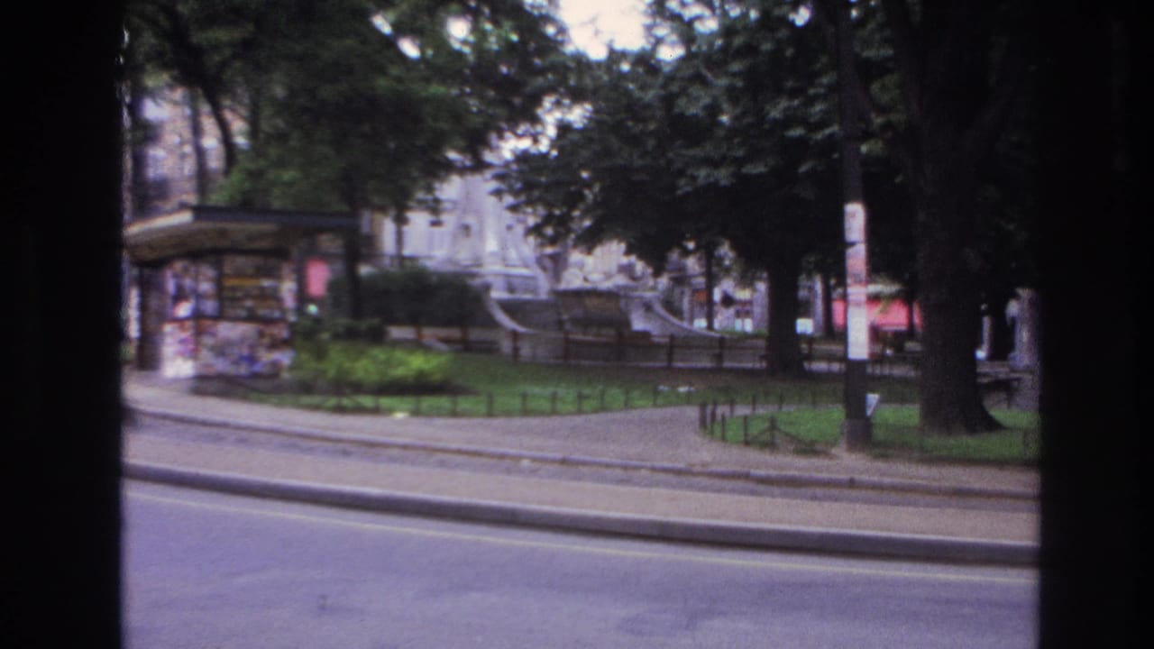 Park entrance area with kiosk, fountain, trees, railings, and street in the foreground.