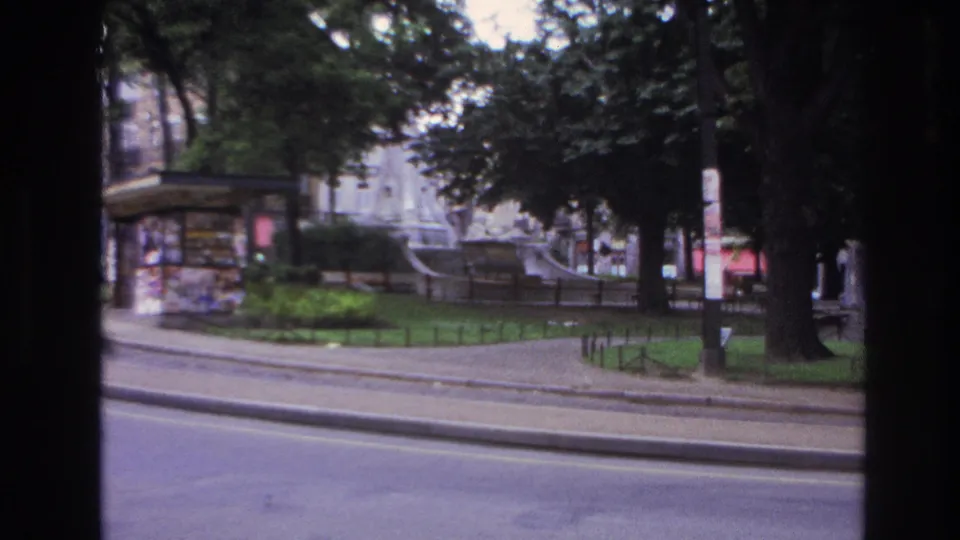 1967 Paris street view from a moving vehicle, with a kiosk near a small park, a fountain behind trees, and apartment buildings beyond.