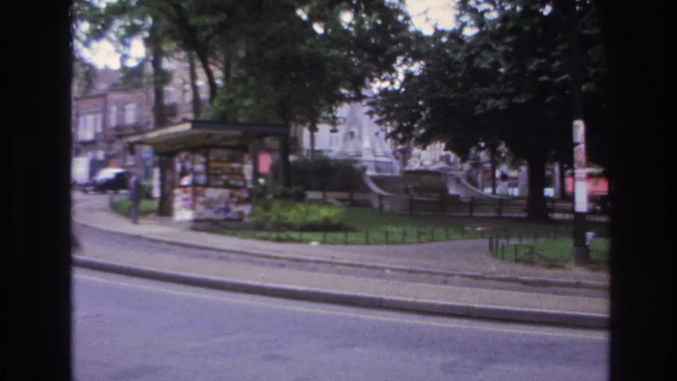 1967 Paris home-movie frame with a person walking near a pavement kiosk, a tree-covered park, road, railings, and older buildings in the background.