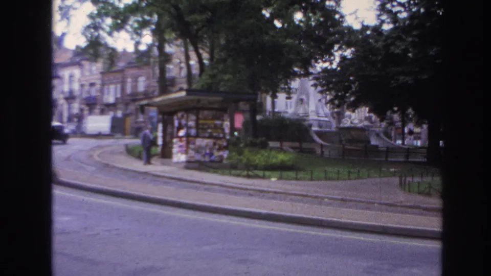 Late-frame 1967 Paris street view showing the kiosk beside the park, a distant pedestrian, trees, fountain structure, railings, and road surface.