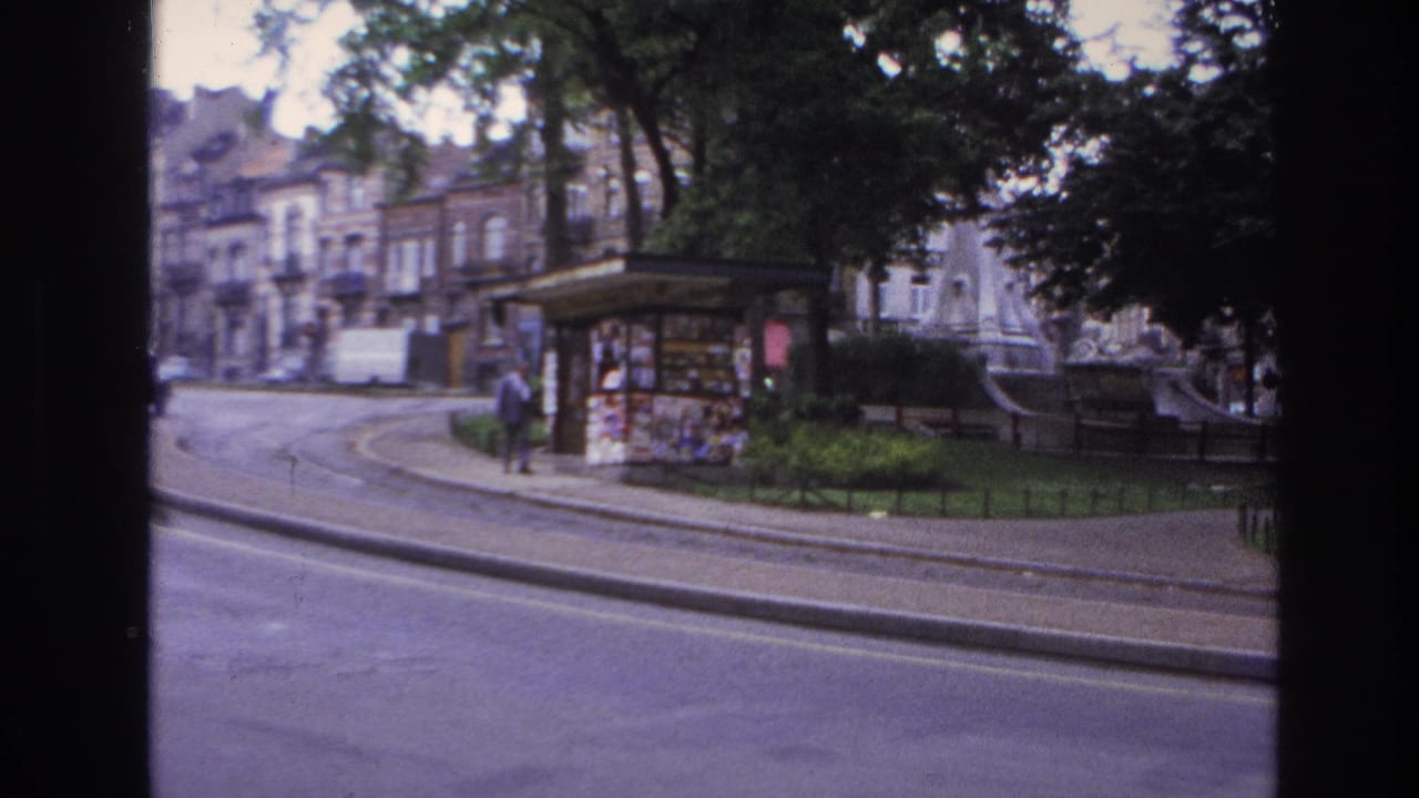 1967 Paris street scene from 8mm footage showing a curved roadway, tram tracks, a kiosk, trees, and a man walking near the sidewalk.