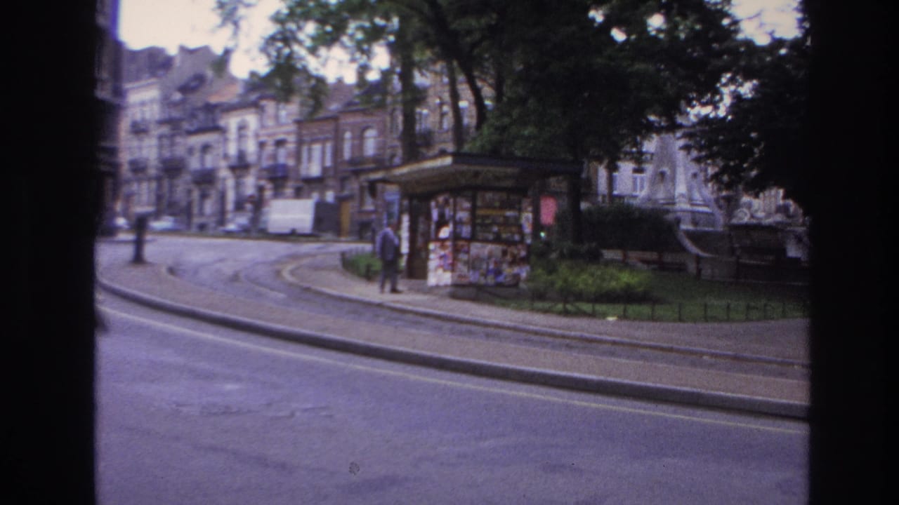 Street corner with kiosk, tram tracks, trees, apartment facades, and a pedestrian near the curb.
