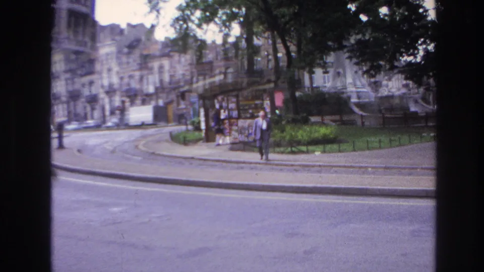 In this 1967 Paris frame, a man in a light-colored suit walks near a sidewalk kiosk beside a curved street with tram tracks and trees.