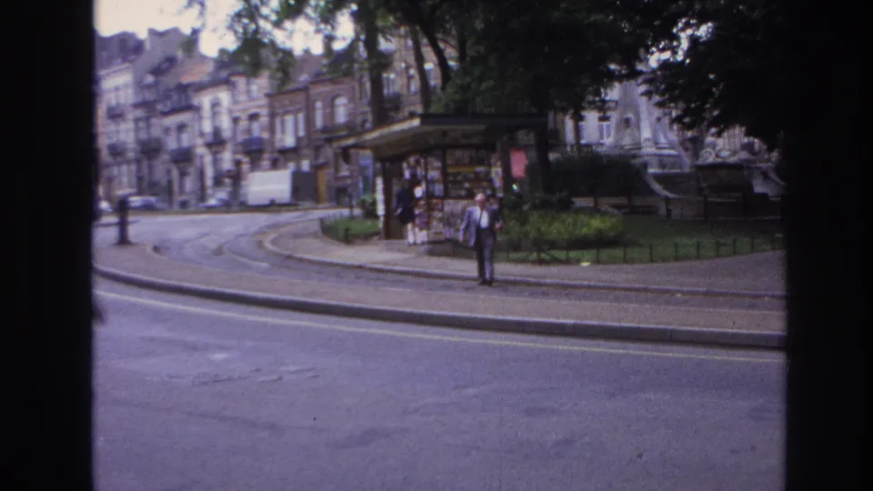 1967 Paris street-side park view with a kiosk, tram tracks, apartment buildings, trees, and an adult man near the curb.