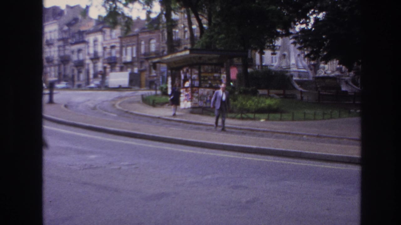 Woman and man walk near a kiosk beside a curved street and park fencing.