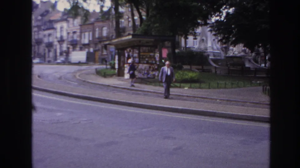 Paris home-movie frame from 1967 showing an adult man walking by tram tracks as another pedestrian moves near a small park kiosk.