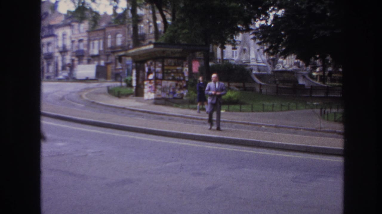 1967 Paris street view from a vehicle window, with a suited man at the curb near a poster-covered kiosk, park railings, trees, and buildings.