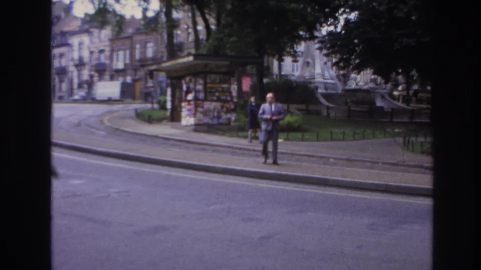 1967 Paris street view from a vehicle window, with a suited man at the curb near a poster-covered kiosk, park railings, trees, and buildings.