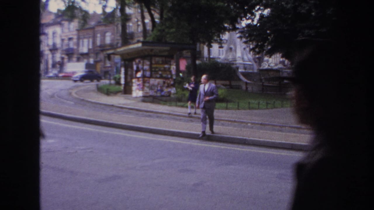 Candid 1967 Paris street frame showing a suited man waiting by the curb while a car moves past near a kiosk, railings, and shaded trees.