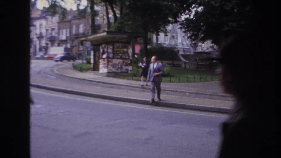Candid 1967 Paris street frame showing a suited man waiting by the curb while a car moves past near a kiosk, railings, and shaded trees.