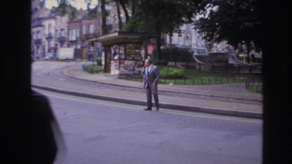1967 Paris home-movie frame of a man in a light suit standing near a curb, seen from inside a vehicle with a kiosk and park edge behind him.