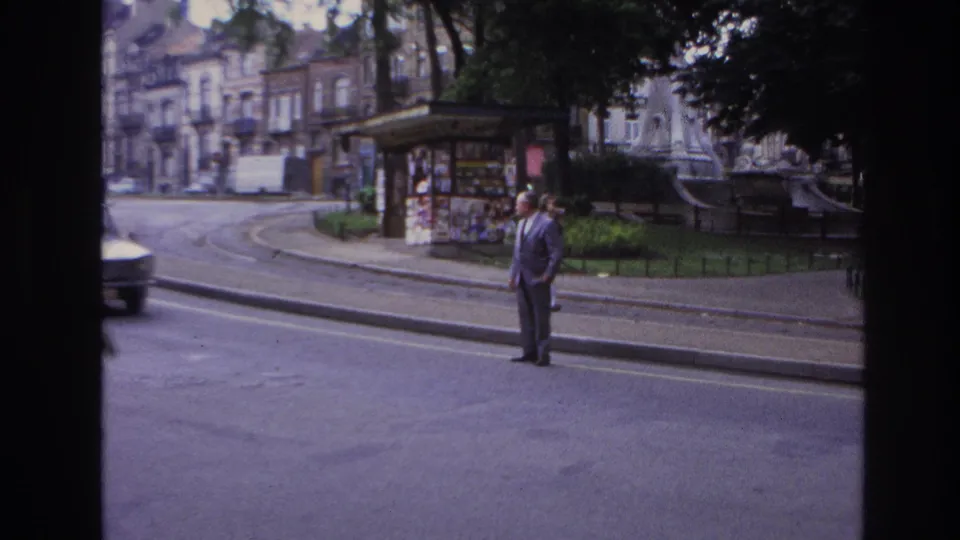 Final 1967 Paris street frame showing a suited pedestrian near the curb as a car enters view, with a kiosk and shaded park edge behind him.