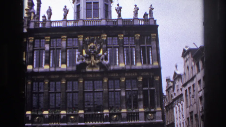 Paris landmark-style facade showing rooftop figures, a central crest, dark window rows, and neighboring stone buildings.