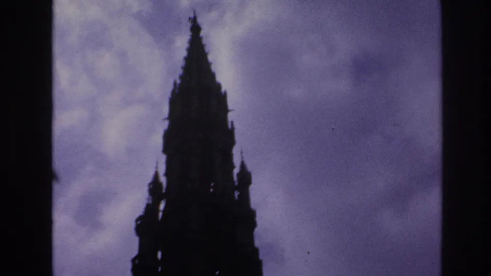 1967 Paris home-movie frame showing a Gothic cathedral spire in dark silhouette against a saturated purple-blue sky.