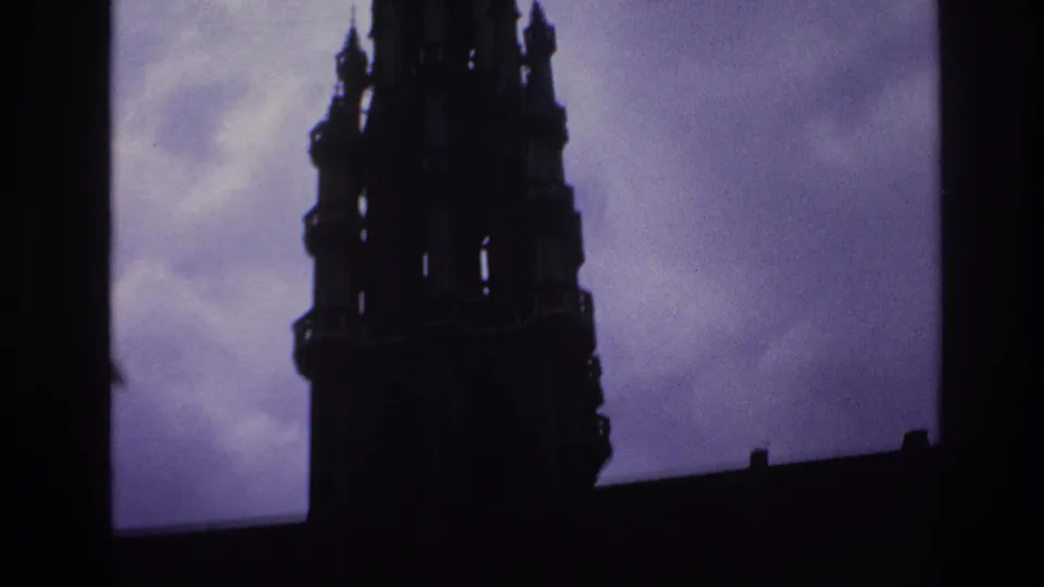1967 Paris home-movie still showing more of the Gothic tower below the spire, with dark stonework against a purple sky.