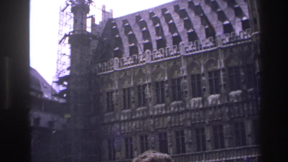 1967 Paris home-movie frame with a tilted view of a historic stone facade, steep roof, scaffolding, dark foreground edge, and a partial head below.