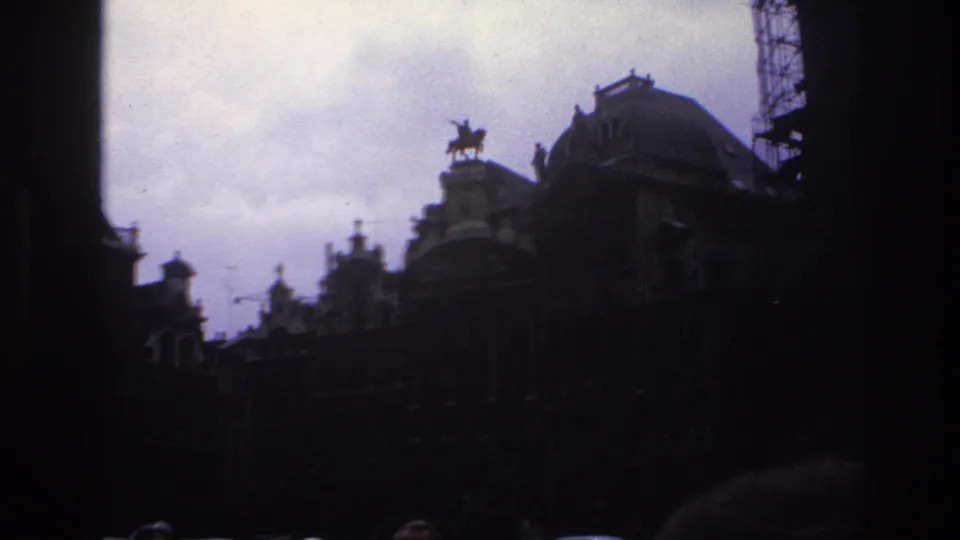 1967 Paris home-movie frame of ornate rooftops in deep shadow, with a domed roof, equestrian statue, scaffolding, and crowd edges.
