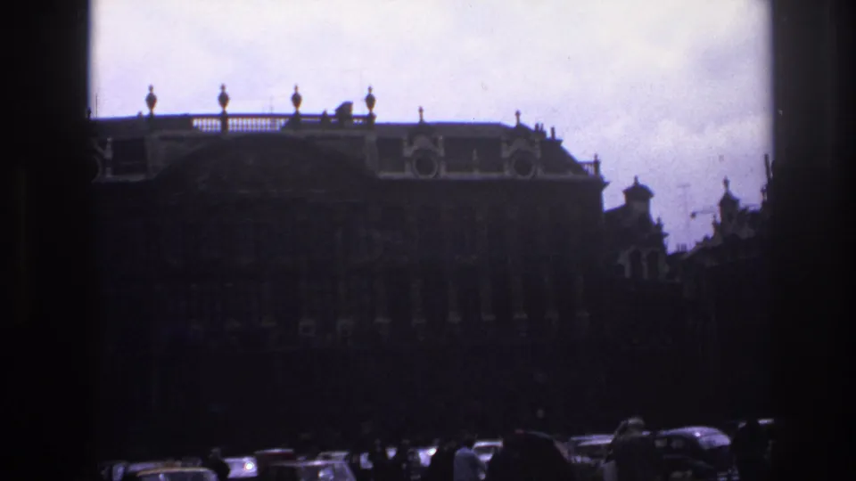 1967 Paris home-movie frame showing a grand historic facade, roof balustrade, cars, and small pedestrian silhouettes under an overcast sky.