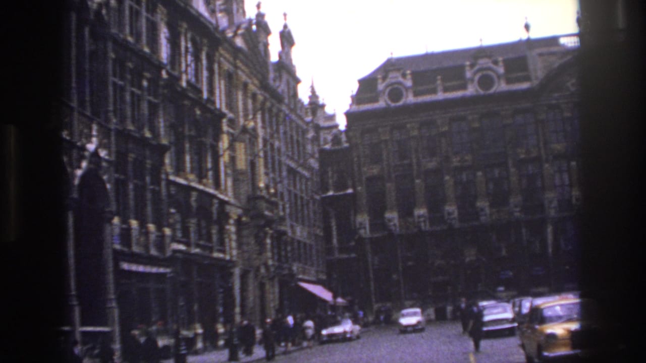 1967 Paris street view with ornate historic facades, pedestrians, and mid-century cars seen from a handheld 8mm film frame.