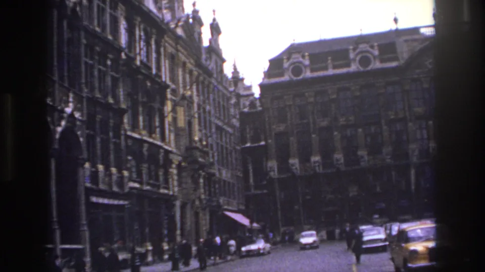1967 Paris street view with ornate historic facades, pedestrians, and mid-century cars seen from a handheld 8mm film frame.