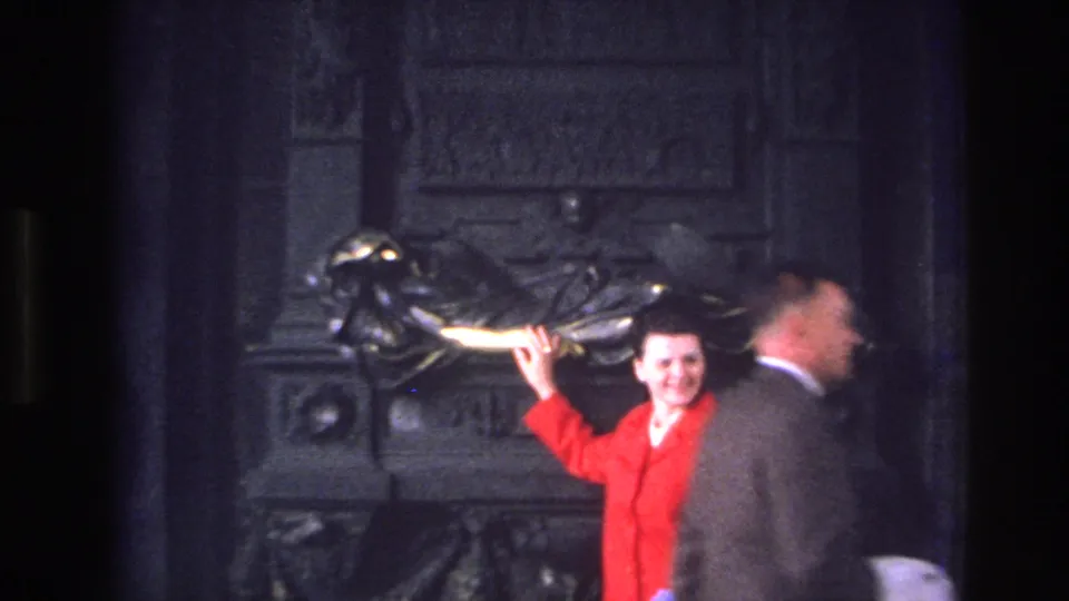 Inside a historic building in Paris, a woman in a bright red coat gestures toward a large sculptural figure while another visitor passes nearby.