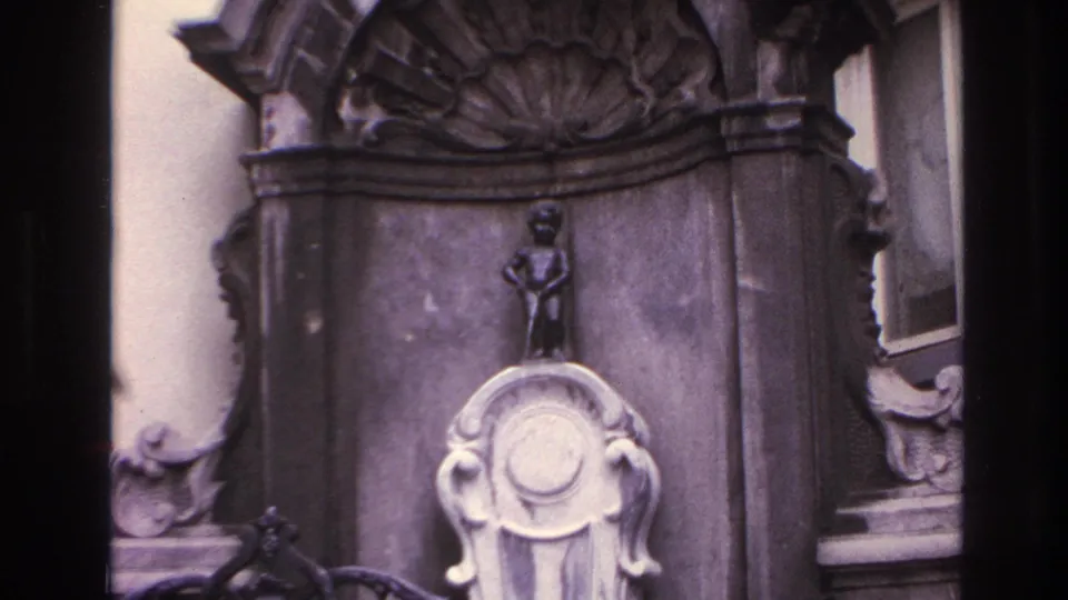 Wider 8mm frame of the ornate stone fountain, showing the statue, basin, carved arch, nearby wall, window edge, and iron railing.