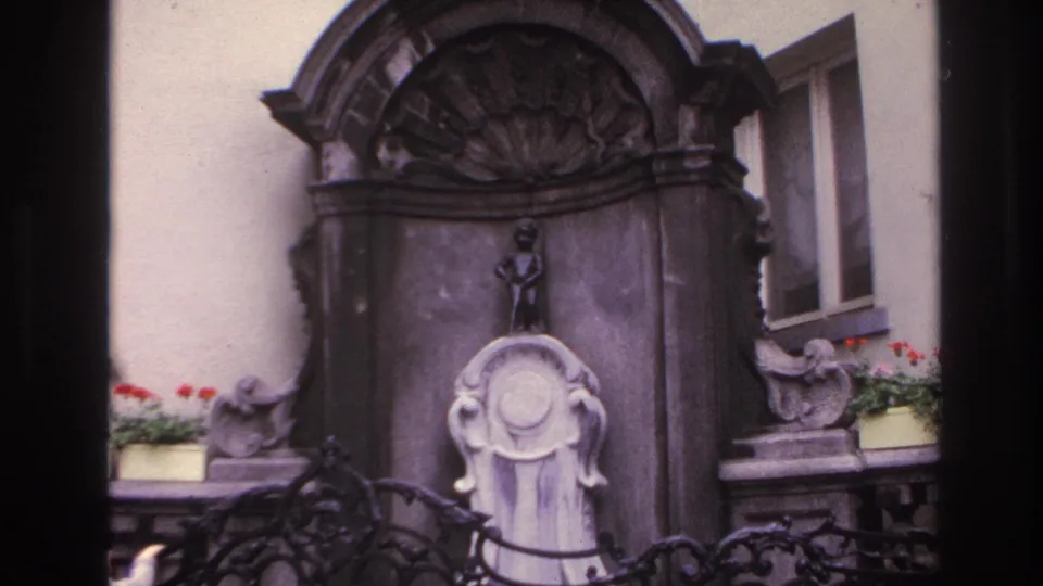 Final wider view of the fountain structure, with the small figure above the basin, iron railing below, and red flowers near a window.