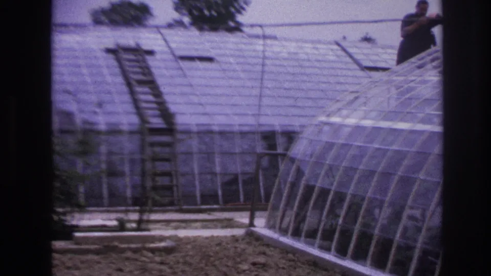 1967 Paris home-movie frame showing a man working above a curved greenhouse roof, with a ladder and glass panes below.