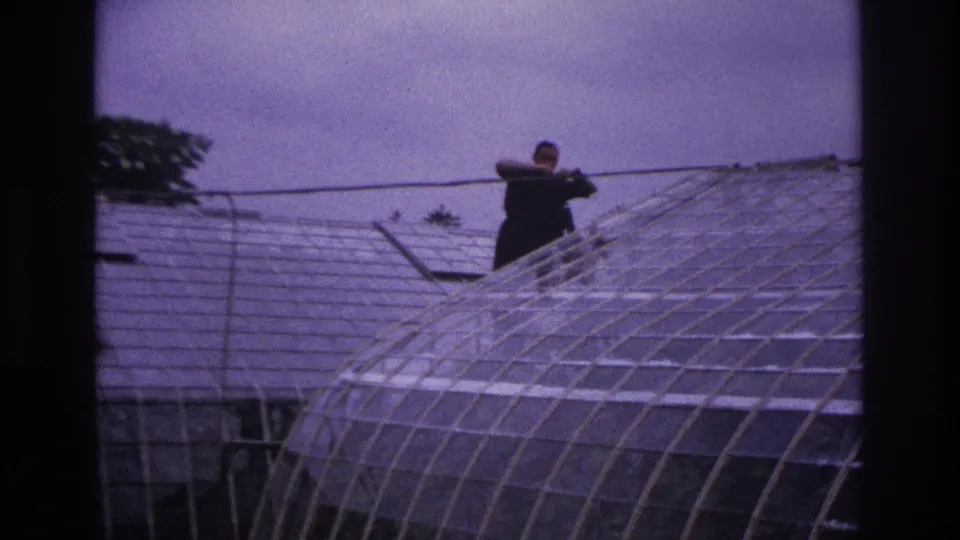 1967 Paris 8mm frame with a distant man using a long pole above the glass panels of a large greenhouse roof.