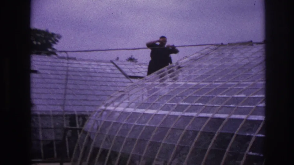 1967 Paris home-movie frame of a man in dark clothing handling a long pole above a curved greenhouse roof.