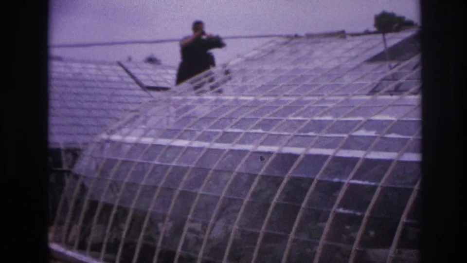 1967 Paris frame showing a distant man using a pole across the roofline of a large arched glass greenhouse.