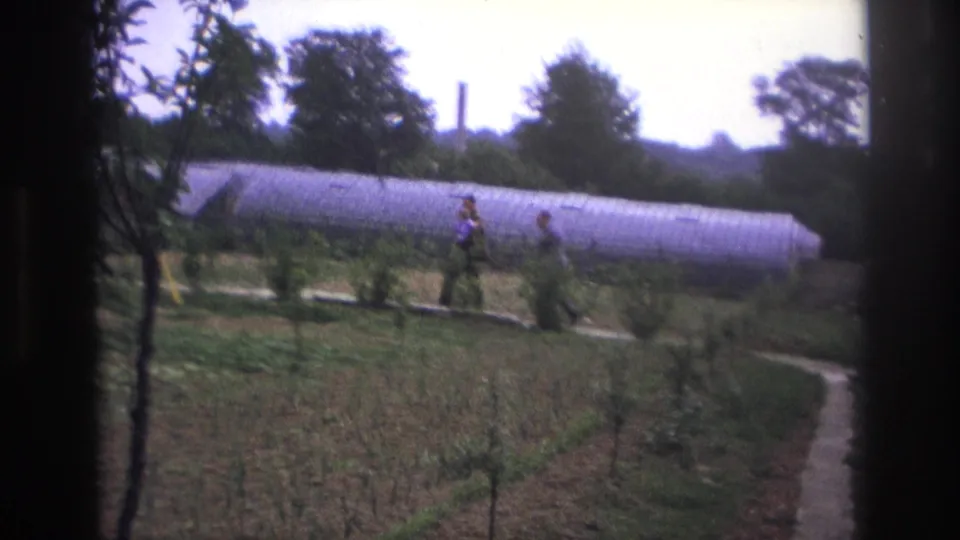 Two men in casual clothing walk through a Paris garden, with a pale arched greenhouse, trees, and planted plots visible in soft 8mm color.